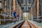 Dwóch rowerzystów jedzie mostem kolejowym.