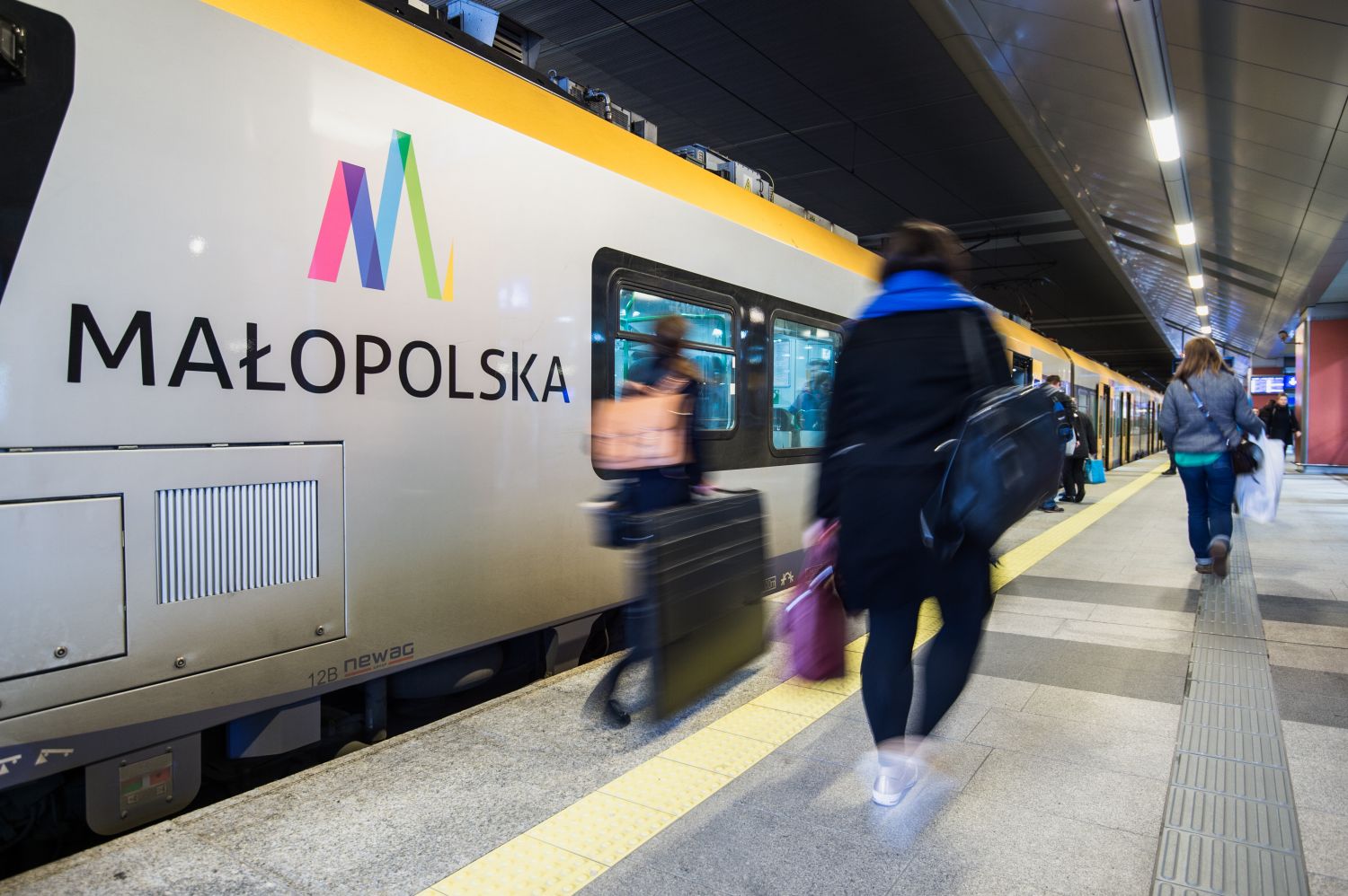 Zdjęcie przedstawia pasażerów wsiadających do pociągu na dworcu kolejowym